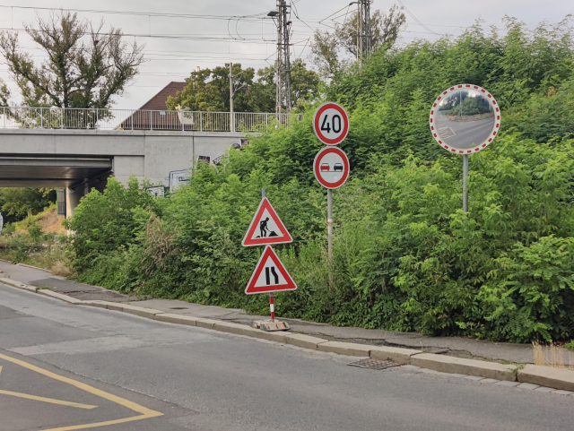 Úzký chodník, který má z jedné strany rušnou silnici, z druhé strany přerostlé křoví a dopravní značku a na něm stojí dočasná značku "pozor zúžení"
