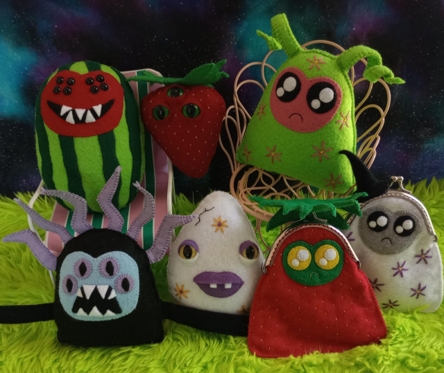 Here's a lovely photo of a plethora of hand-sewn fabric art creatures. There's a watermelon creature with a red face, eight black seed-like eyes, and a wide mouth with five sharp teeth. There's a white ghost trinket bag covered with purple embroidered flowers. She has a gray face, sparkly black eyes and a little frown. There's a strawberry creature with three bulbous green eyes. There's a watermelon ghost, a strawberry ghost bag, and a black creature with sharp teeth. There's a white egg with yellow embroidered flowers and purple eyeshadow and purple lips. All the creatures are sitting on green fur and there's a galaxy print fabric in the background. The watermelon creature and the watermelon ghost are sitting on metal doll chairs.
