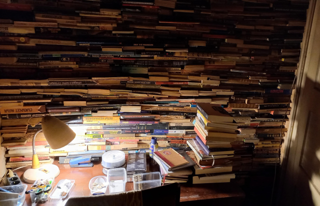 A wall of stacked books over a desk with a lamp in the late evening. #bookstodon