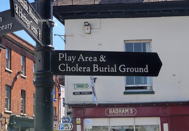 An old fashioned wrought iron sign with an arrow pointing to "Play Area & Cholera Burial Ground"