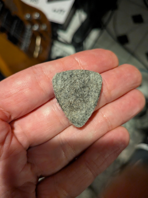 A gray felt pick, held in an outstretched hand. Blurred and in the background is an electric guitar on a stand.