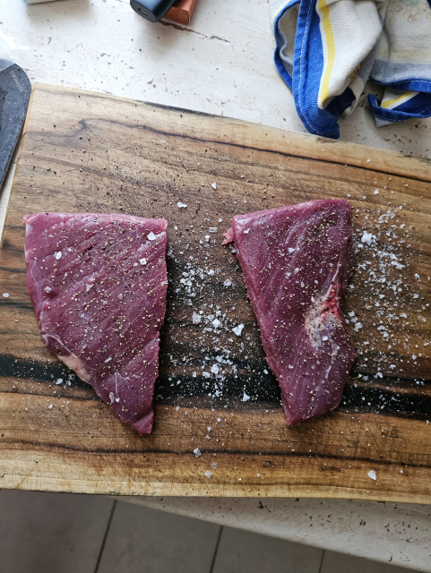 fotka 1: na drevenej doske rozkrojený stejk na dve časti, okorenený a osolený, korenie je aj navôkol, okolo dosky utierka iqos a tak podobne