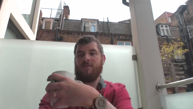 From my laptop's webcam, an image of me holding a coffee mug. I'm wearing my Bradley timepiece on my left wrist, a maroon polo shirt and a set of earbuds.  The background consists of a frosted glass barrier or wall, and behind it, a brick building with several windows can be seen. There's a tree with yellowing leaves on the right-hand side. The sky appears bright and slightly overcast, and the top of a building structure is visible in the upper part of the image, suggesting a cityscape 