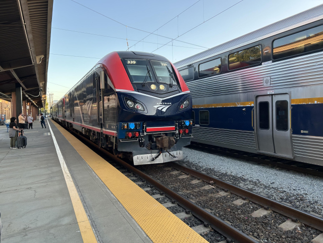 A red and dark blue Amtrak locomotive pulling a long train into San Jose station.
