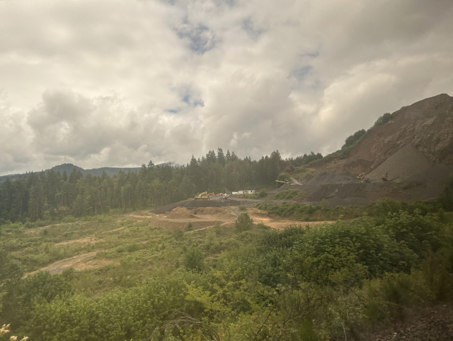 View from the train. A grassy area with dense trees in the background. A hill on the right. Patchy clouds covering most of the sky.