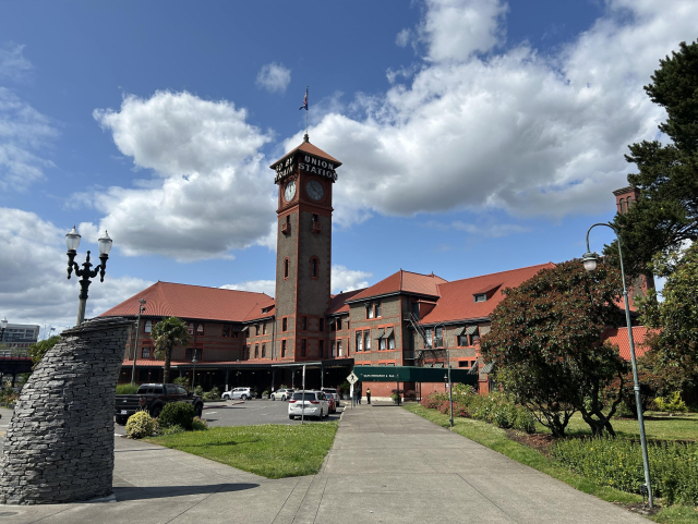 The Portland train station from the street side. It’s a brown building with red tile roof and a clock tower which says “go by train” on one side and “union station” on another side. Green grass and other plants all around in front of the building. Big poofy clouds in the sky, but lots of blue showing.