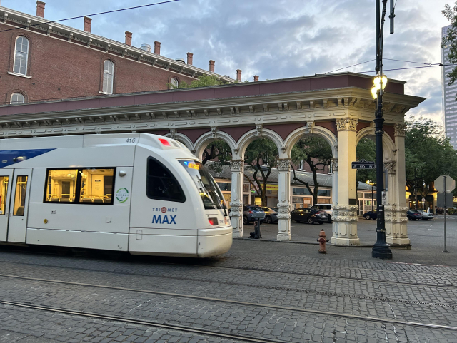The rounded end of a white MAX light rail train passing an ornate colonnade and brick building at dusk. The ground has two sets of tracks and is paved with cobblestones.