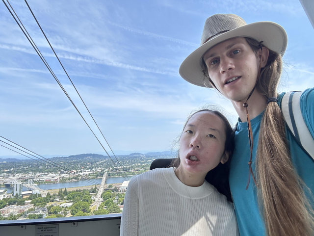 Aubrie and Peter at the top of the aerial tram, with cables running down on the left across the wispy cloud sky. Peter is wearing a tan hat.