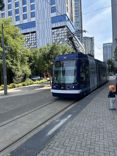Teal-fronted NS streetcar approaching a station. Trees line the other side of the street. Tall building behind.