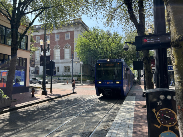 A blue (in paint color) MAX blue line light rail vehicle approaching a station in downtown. A brick building in the background. Lots of trees around.