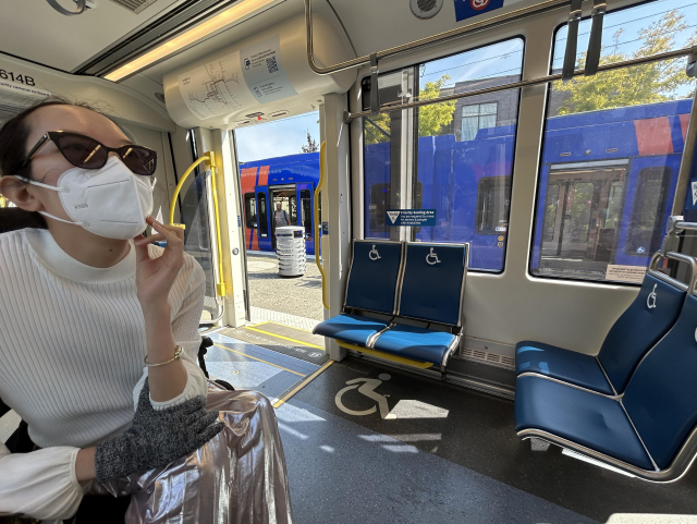 Accessible seating area of MAX train, with wheelchair symbols everywhere. Aubrie sits in her power wheelchair at the left, wearing a shiny silver skirt and white shirt. The train door is open and a blue train is sitting at the other side of the island platform.