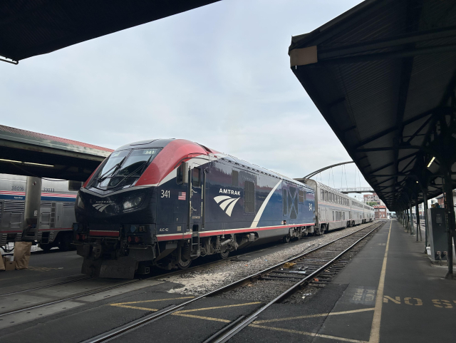 An Amtrak passenger train sitting at Portland station, with the locomotive close up and several double-decker passenger cars behind it. Roofs are over the platforms on each side of the train. An arch passenger bridge goes over the tracks near the end of the train. The whole sky is grey.