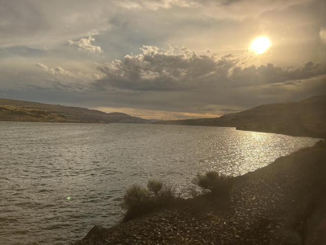 The Columbia river reflecting a bright, low sun. The sky is filled with soft and detailed clouds. The banks of the river cut across the foreground and recede into low mountains on the horizon.