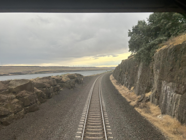 A view out the back of the train. The track extends away and curves to the right around a cliff face topped with trees. The track is bordered by ballast that fades from light to dark gray as it gets further from the rails. On the left are some low rocks, and beyond that, the Columbia river. The picture has a dynamic angled quality to it, with a soft gray and yellow sky taking up the upper left side, and the rocky land and curving track cutting across the lower and right side.