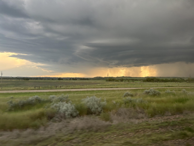 A dramatic sky close to sunset over a green landscape. Dark, heavy clouds swirl in the air, and in the distance, a flame-like mist glows with golden light.