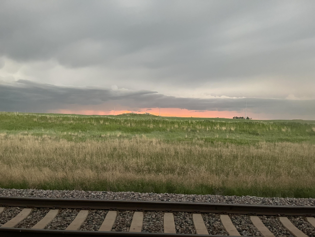 A clouded dusk over grassy plains. A thin slice of pink light separates the land and sky. Tracks span the lower edge of the picture.