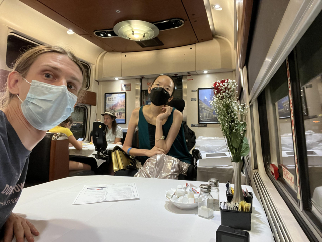 Peter taking a selphie of Peter and Aubrie sitting in the dining car across from each other at a table. The table has a white tablecloth and some fake flowers, half and half, and condiments at the edge near the window. Peter wears a blue mask and Aubrie wears a black mask and a very shiny skirt. A somewhat fancy light is on the ceiling. Right behind Aubrie are two white trash receptacles.