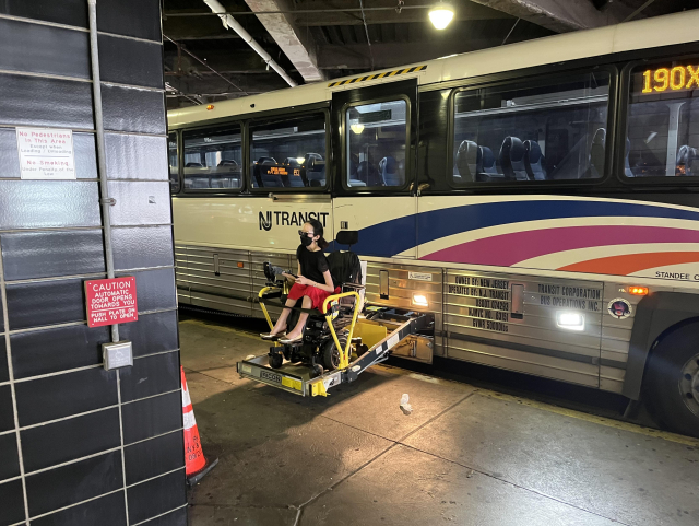 A New Jersey Transit bus parked in the Port Authority Bus Terminal. Aubrie is in her power wheelchair on the lift coming out of the side of the bus. The bus is parked in such a way that the lift is aligned with a giant structural column of the building, constraining the way Aubrie can roll off the lift when it reaches the ground.