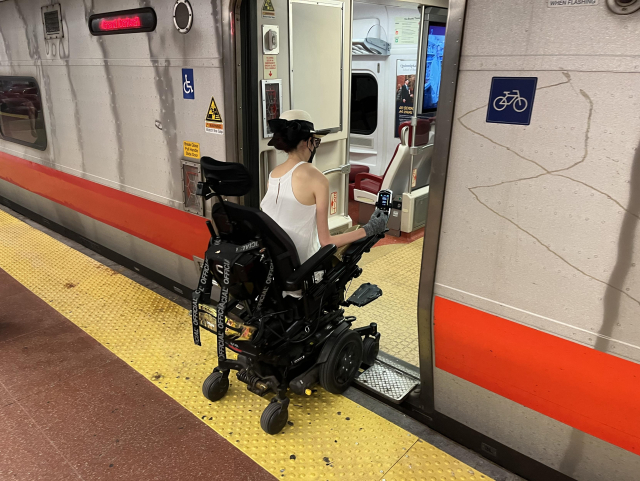 Aubrie in her power wheelchair rolling onto a Metro North train. A little metal plate extends from the train floor toward the yellow bumpy platform edge.
