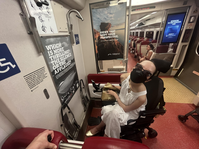 Aubrie sitting in the windowless wheelchair area right next to the doors of the train. Seats are folded up on either side of her.