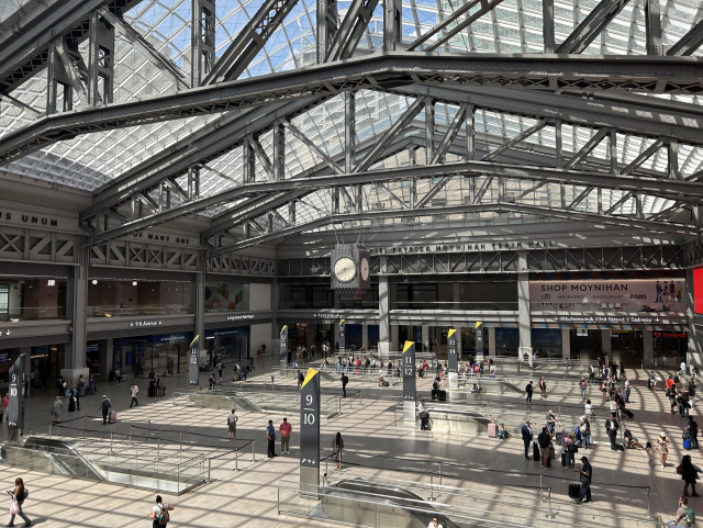 A view of Moynihan Train Hall from the lounge on the second floor, overlooking the main floor with many people. Escalators puncture the otherwise smooth, uninterupted floor. The ceiling is made of many glass panes, with intricate metal beams crossing it in archlike structures. A clock hangs in the center of the ceiling, showing the time is 2: 38.