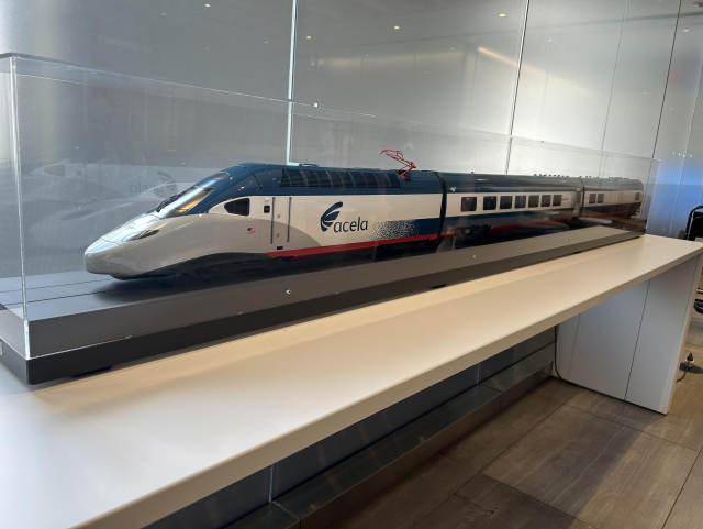 A model of the new Amtrak Acela high speed train inside a clear case sitting on a long table against a wall. It has a sleek, pointed nose and three cars, including the front one. The train is dark blue along the bottom amd top and white along most of the sides. A blue gradient graphic runs along the bottom of the white section. It says acela in a few places.