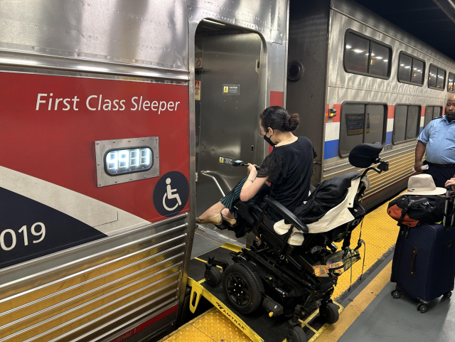 Aubrie, in her power wheelchair, rolling up a black and yellow bridge plate from the platform into a train. It’s a shiny, silver, single-level train with tall, red stripe running along its side, on which is white text that says, “First class sleeper”. A blue triangular section below the red stripe shows the last three digits of a number, which are 019. A ♿️ is next to the door. An Amtrak employee is partially in the photo on the right. A suitcase with a hat resting on top of it sits on the platform at the right.