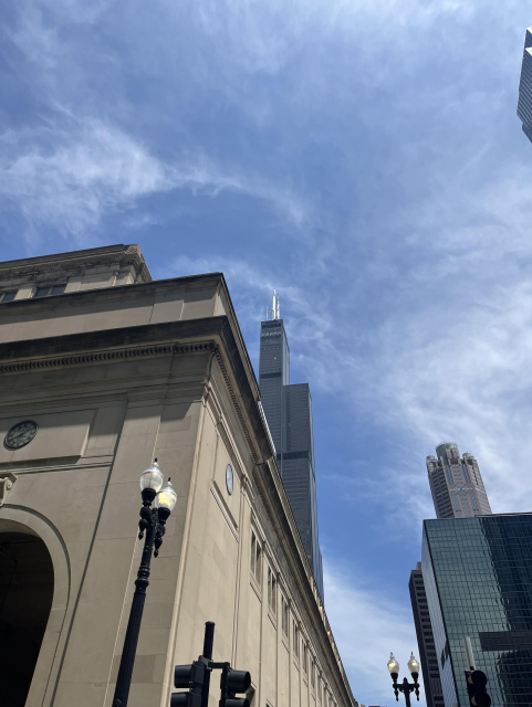 Looking up toward the southwest corner of the ornate, stone Chicago Union Station building, behind which the Willis Tower juts into the sky, with its dark facade and white antennae. Wispy, white clouds lightly cover the sky.