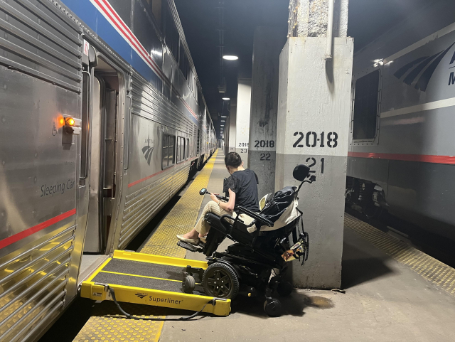 Aubrie in her power wheelchair attempting to roll onto a black and yellow ramp from the underground platform into a double-decker Amtrak Superliner train car. A large concrete column is right at the bottom of the ramp, so her chair is wedged between the column and one side of the bottom of the ramp. She looks down toward the column, trying to maneuver.