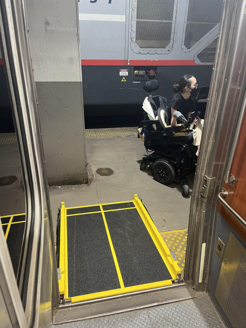A view from just inside the doorway of the train, showing the ramp, which is halfway blocked by the concrete column on the platform. Aubrie sits in her power wheelchair on the platform. Her chair clearly won’t fit past the column onto the ramp.