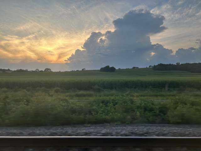 A view out the train window at sunset in western Iowa. A rail is along the bottom. Above that, tall, soft, green grass extends into the distance. A few trees in the distance. In the sky, a dark, billowing cloud covers the right center of the photo, getting narrower near the top, where its edges are backlit by the sun. Behind it, the sky is covered in layers of pillowy clouds at the bottom, and more wispy clouds at the top, which are all illuminated, displaying a range of colors from yellow to orange to dark reddish and purple. The contrast between the dark cloud and lighter background clouds is stark.