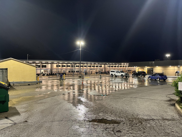 A view across the parking lot at Omaha train station after midnight. The ground is incredibly wet, but it’s not currently raining. A handful of cars are parked. Some double-decker Superliner train cars sit at the platform spanning the width of the photo, reflected down onto the soaked asphalt. A small tan building is at the left next to the parking lot. A larger tan building with a dark, blockular roof is at the right. Lights on poles in the parking lot cause lens flares across the photo.
