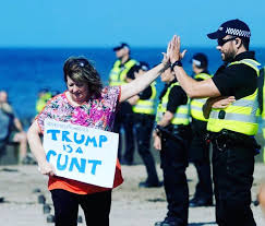 Late Scottish comedian Janey Godley high fiving a Police Scotland officer while carrying her iconic TRUMP IS A CUNT sign as she walks on the beach at Turnberry in South Ayrshire, Scotland during Trump's visit to his golf course in summer 2016 