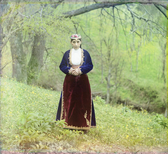 an Armenian woman posing in a dark red skirt, a white shirt, and a blue jacket, wearing a multi-coloured hat, on a hillside against a backdrop of trees. her hands are clasped in front of her stomach.