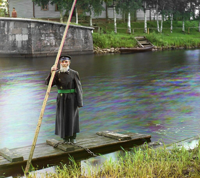 an elderly, bearded man in a formal uniform standing on a raft holding a long punting pole. the raft floats on a river but its chained to the bank. the man's coat is long and grey, buttoned on the right side with a green belt. in this one you can see the effects of taking three separate photographs to composite them; while most things in frame are static and thus line up perfectly, the river is in constant movement, and so the water is separated into clouds of all three colours and looks somewhat oily.
