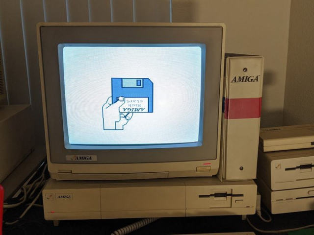 a beige desktop computer with a built-in floppy drive. on top is a beige monitor with a grey bezel displaying a graphic of a hand holding a blue floppy disk with a label that (upside down) says "Amiga Kickstart." Next to the monitor is a beige binder with a red stripe across it and the Amiga logo.
