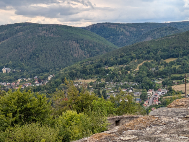 Eine bewaldete Berglandschaft, durchzogen von einem tiefen Taleinschnitt. Darin eingebettet liegt der Ort Bad Blankenburg. Im Hintergrund bewölkter Himmel, der den Schein der Sonne ahnen läßt. Rechts unten im Vordergrund eine breite verwitterte Steinmauer.