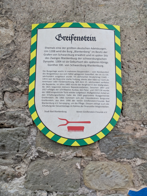 An einer Mauer ist eine wappenartige Informationstafel angebracht.