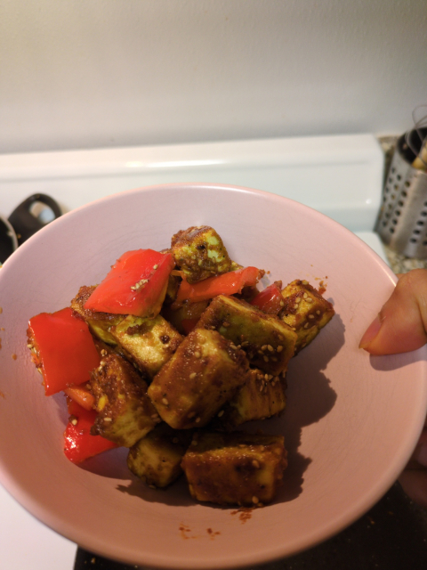 Crispy yellowish tofu seasoned heavily with an orange-brown coating and sesame seeds, with some red bell pepper chunks in a pink bowl.