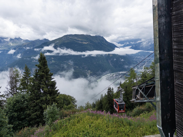 Seilbahn aus dem Tal kommend mit einer roten Kabine, im Hintergrund wolkenverhangene Berge
