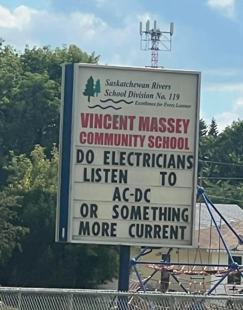 The image shows a sign for Vincent Massey Community School in front of a backdrop of trees and a cloudy sky. The sign is rectangular with a light background and dark blue borders. At the top of the sign, there is the logo for Saskatchewan Rivers School Division No. 119, including a stylized image of trees and waves. The words "Excellence for Every Learner" are written below the logo. The main text of the sign reads "VINCENT MASSEY COMMUNITY SCHOOL" in large, red lettering. Below this is a question: "DO ELECTRICIANS LISTEN TO AC-DC OR SOMETHING MORE CURRENT" in black letters. The sign appears to be made of individual letters that can be changed. In the background, there are lush green trees and a telephone tower. A chain-link fence is visible in the foreground. A playground structure can be seen on the right side of the image. The overall impression is a playful, lighthearted message on the school sign. The color palette is dominated by greens, blues, and whites. The composition is centered on the sign, which occupies a significant portion of the frame.