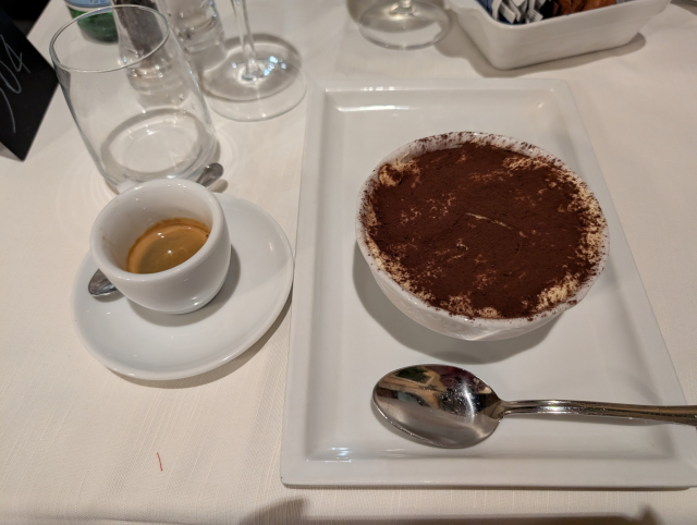 A bowl of tiramisu with a spoon on the right with a decaf espresso on the left for scale