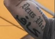 A close up shot of current US Defense Secretary Pete Hegseth flexing his bicep. A tattoo of the words "Deus Vult" ("god wills it") is visible. Underneath is another tattoo of the word "infidel" in Arabic.