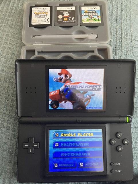 A Nintendo DS showing the Mario Kart DS main menu; the console is resting on a cartridge container box, showing a stylus and three additional DS games: Pokémon White, Grand Theft Auto Chinatown, and New Super Mario Bros