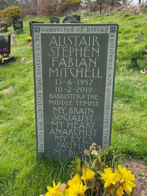 Gravestone inscribed: Alistair Stephen Fabian Mitchell. 13-6-1957, 10-2-2019. Barrister of the Middle Temple. My brain, socialist. My heart, anarchist. My eyes, pacifist. My blood, revolutionary. 

Around the edge, the legend: The only man in British legal history to be convicted of biting a policeman with someone else’s teeth.
