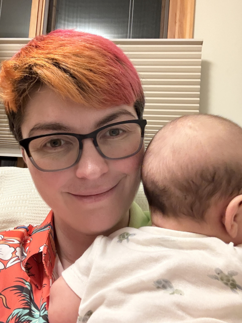 A picture of a smiling woman with pink and orange hair, wearing an orange Hawaiian shirt. She is holding a baby who is facing away from the camera. He is wearing a white onesie with little green turtles on it.