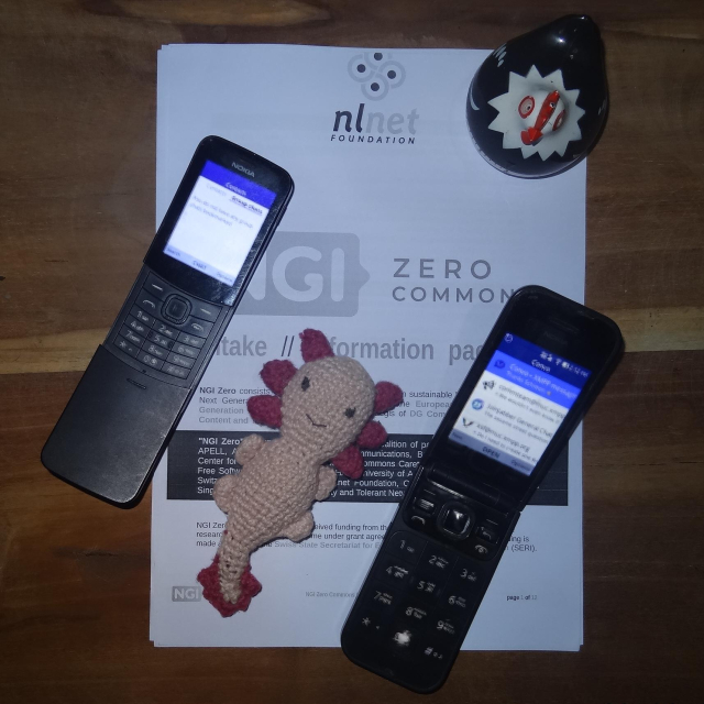 A printout of an NGI Zero Commons fund information document, with a few things placed on top. Diagonally from top left to bottom right are: A Nokia 8110 "Bananaphone", an amigurumi (crochet) axolotl, and a Nokia 2720 Flip phone. On the top right corner of the paper is a round clockwork hen named clock-a-doodle-doo.

Both phones are running the Convo app: one with an empty group chat list, and another with a list of conversations. If you look closely and get past the pixellation and blurred-ness, you might recognise group chats with icons for: Convo, XSF Communications Team, XSF Discussion, and JoinJabber General Chat.