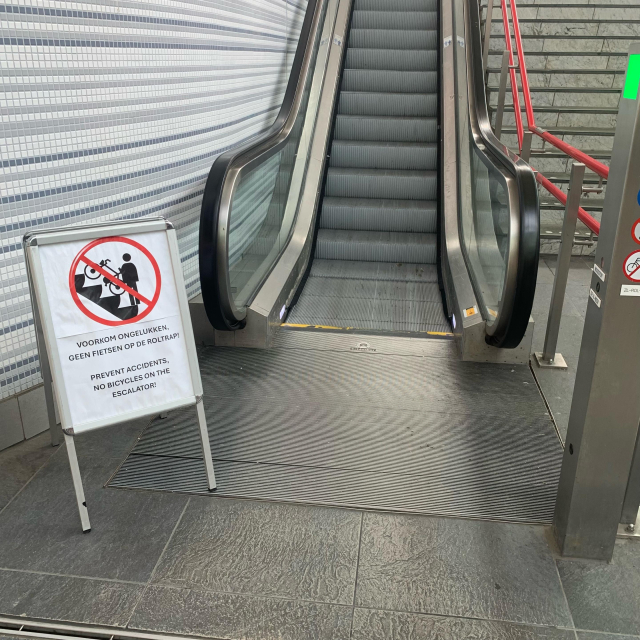 Small sign next to an escalator stating in two languages and a pictogram that using an escalator with a bicycle is forbidden.
