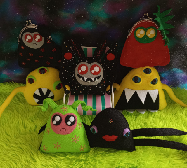 Here's a lovely photo of a plethora of my fabric art creatures. There's a green fur floor and a galaxy print fabric background in the pic. Here are some of the creatures: a watermelon ghost with a green body and pink face and twinkly black eyes and a little frown. There's a black crablike creature with wide set blue eyes and purple eyeshadow. She has red lips, four tentacle-like arms and an embroidered rainbow flower on her forehead. There's a black ghost bag covered with red embroidered flowers. There's a watermelon ghost bag. There are two yellow creatures. Each has three eyes, two tentacles, and mouths full of sharp teeth. There's a cosmic moth sitting on a little chair. The moth is black with a gray face, twinkly red eyes, and a wide grin full of sharp teeth. The moth is covered with hand-embroidered stars and planets of various sizes and colors.