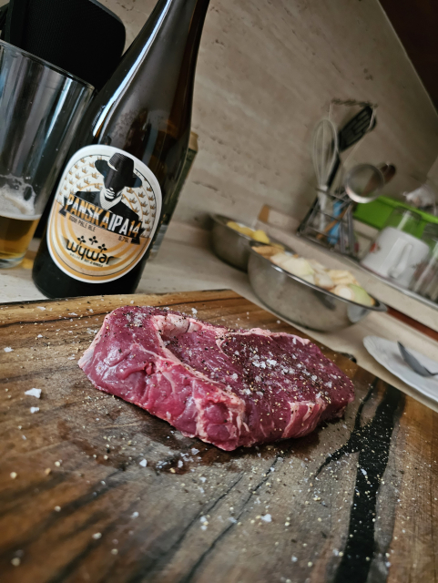 v popredí na drevenej doske stejk rib eye a v pozadí zelenina a pivo 